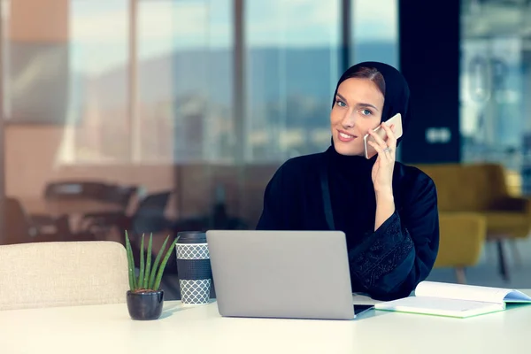 Happy muslim businesswoman in hijab at office workplace. Smiling Arabic woman working on laptop and talking on smartphone — Stock Photo, Image