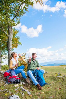 Genç çift dağlarda seyahat ederken kayanın üzerinde dinlenirken su içiyor.