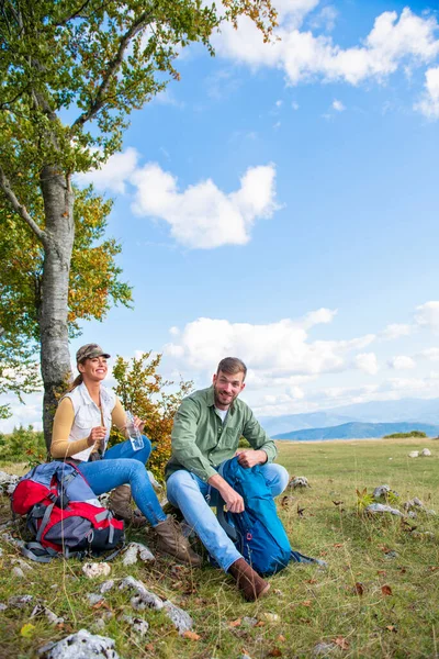 Genç çift dağlarda seyahat ederken kayanın üzerinde dinlenirken su içiyor.