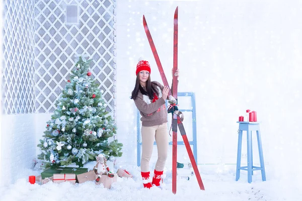 Mavi gözlü, rahat örülü kırmızı atkı ve şapkalı genç bir esmer Noel ağacını süslüyor ve Noel ağacı ile Noel Baba 2020 2022 Noel Baba 'sından hediyeler alıyor.