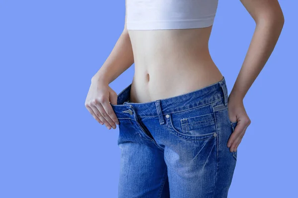 Het concept van dieet en gewichtsverlies. Een vrouw in grote jeans op een blauwe achtergrond toont haar slanke buik. Liposuctie. — Stockfoto