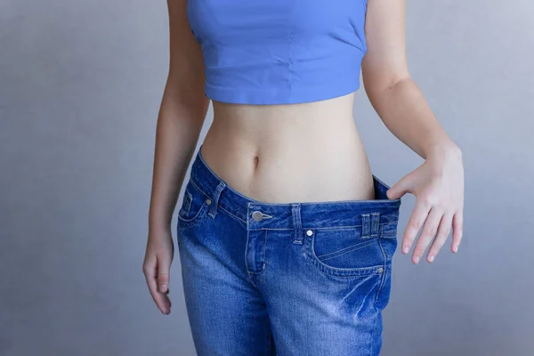 Het concept van dieet en gewichtsverlies. Een vrouw in grote jeans op een blauwe achtergrond toont haar slanke buik. Liposuctie. — Stockfoto