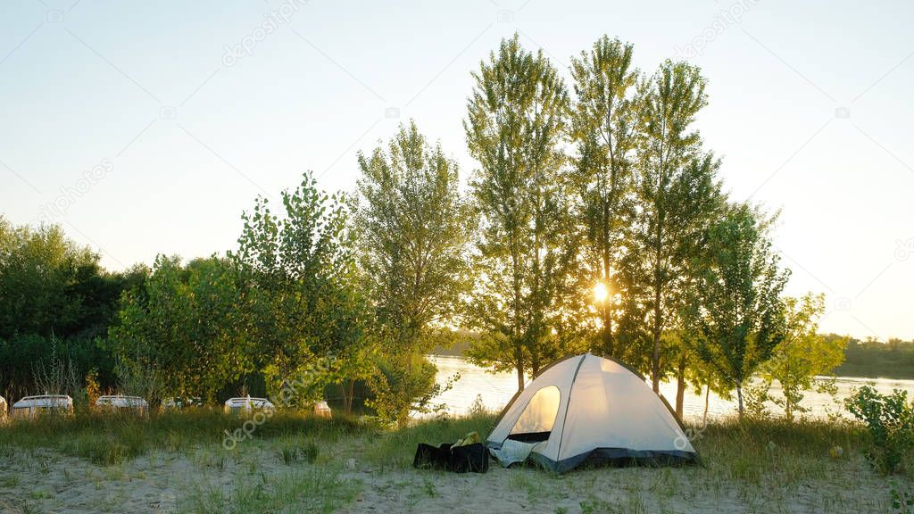 tourist tent stands on the sandy bank of the river in the summer evening is illuminated by the sun through the trees
