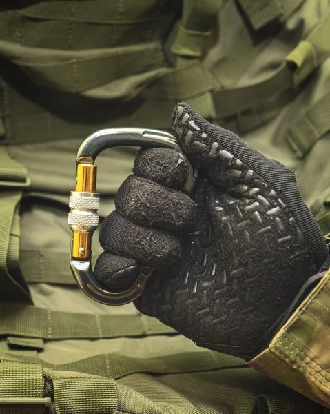 a hand in a black army glove holds an aluminum climbing carabiner against the background of a green military backpack