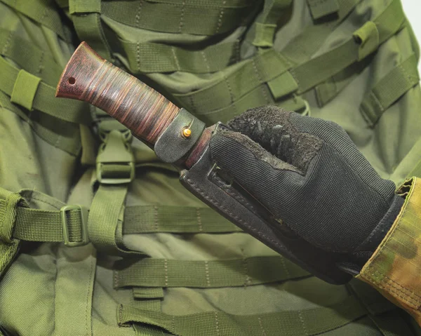 a hand in a black army glove holds a knife against the background of a green military backpack