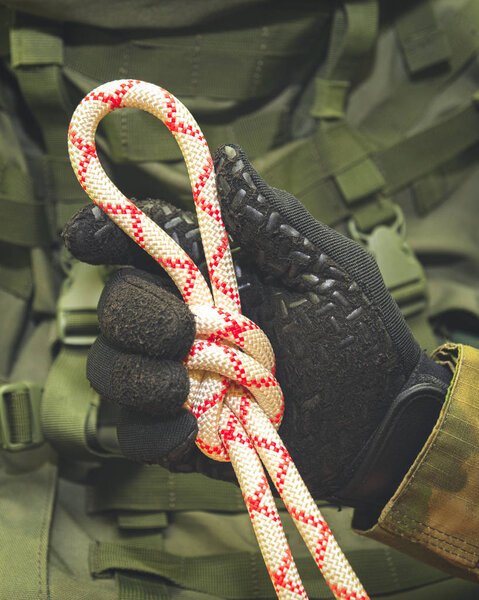 рука в черной перчатке держит альпинистскую веревку, завязанную в узел "восемь" на фоне военного рюкзака

