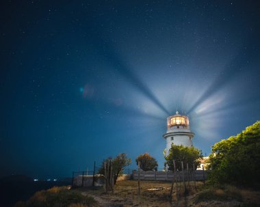 Feodosia'da beyaz deniz feneri, Kırım'da Karadeniz'de gece gökyüzünün altında ışık ışınları ile