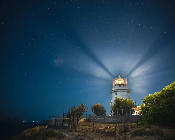 Feodosia'da beyaz deniz feneri, Kırım'da Karadeniz'de gece gökyüzünün altında ışık ışınları ile