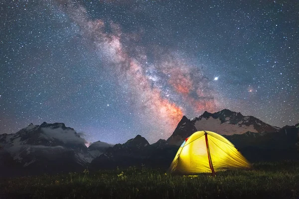 Dağ zirvelerinin ve gece gökyüzündeki Samanyolu 'nun arka planında bir çayırda turist sarı çadırı