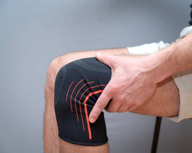 Erkek eli, dizinde siyah elastik bandajla hafif bir arka planda durur.