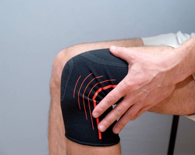 Erkek eli, dizinde siyah elastik bandajla hafif bir arka planda durur.