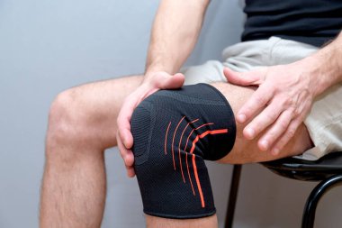 Erkek eli, dizinde siyah elastik bandajla hafif bir arka planda durur.