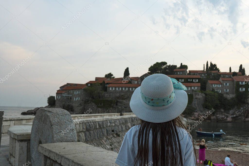 Girl in a hat on the shore of the waterfront. Islet Sveti Stefan and resort in Montenegro. Budva Riviera. Balkans, Adriatic Sea, Europe.
