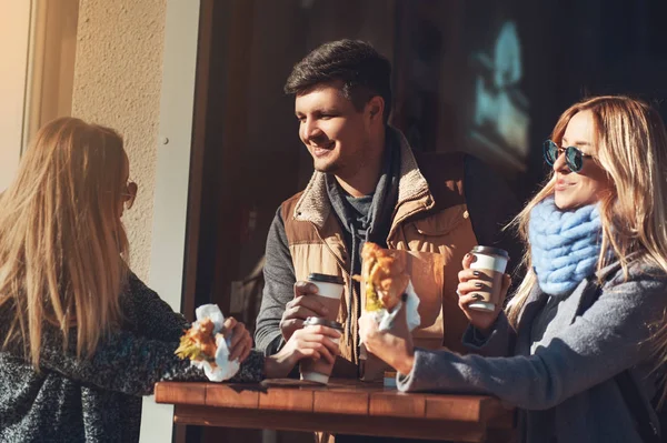 Passer du temps avec des amis. Groupe de jeunes gens joyeux parlant entre eux tout en mangeant croissant et sandwichs et en buvant du café tout en restant debout à l'extérieur . — Photo