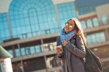 Hava bugün yürüyüş için harika. Kahve fincanı tutarak ve açık havada yüzündeki gülümseme ile yürüyüş güzel sarışın genç kadın.