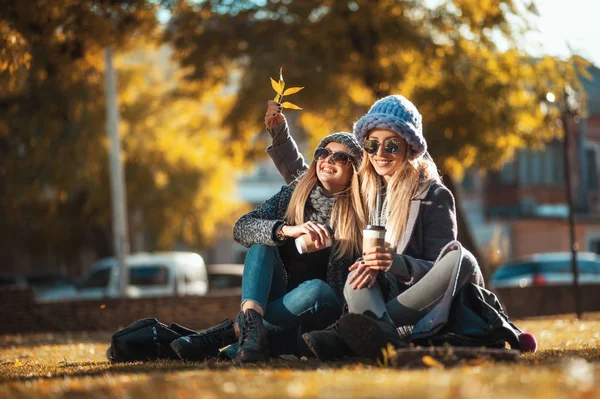 Δύο όμορφες γυναίκες χαρούμενος κάθεται στο πάρκο στο χόρτο πίνει καφέ και γελώντας. Έννοια φιλίας, συναδέλφωσης, ευτυχία. — Φωτογραφία Αρχείου