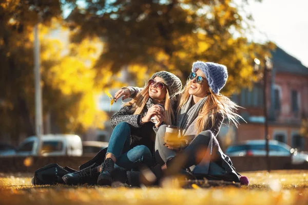 Δύο όμορφες γυναίκες χαρούμενος κάθεται στο πάρκο στο χόρτο πίνει καφέ και γελώντας. Έννοια φιλίας, συναδέλφωσης, ευτυχία. — Φωτογραφία Αρχείου