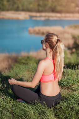 Eğitimden sonra rahatlatıcı. Otların arasında oturan ve güzel doğal nehir arka plan üzerinde meditasyon yaparken şık eşofman çekici genç kız. Kadın Yoga outsid