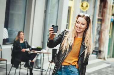 Kahve götürün. Kahve fincanı tutarak ve cadde boyunca yürürken gülümseyen siyah şık kıyafetler giyen güzel genç kentsel kadın. Öğrencinin çalışma sonra kahve molası. Moda yaşam tarzı