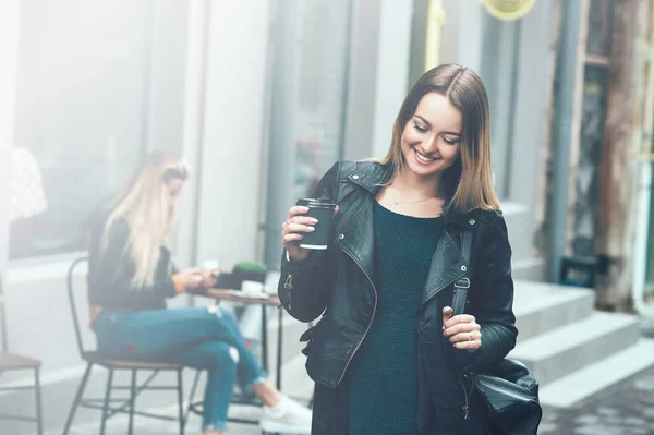 Tira o café. Bela jovem mulher urbana vestindo roupas elegantes pretas segurando xícara de café e sorrindo enquanto caminha pela rua. Pausa de café do estudante após o estudo . — Fotografia de Stock