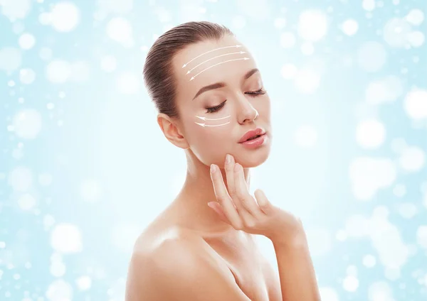 Güzel genç kız mavi arka plan ve kar okları kaldırma ile yüzünü dokunaklı. Plastik cerrahi, facelift ve gençleştirme kavramı. Moda, tatil, kozmetik, 