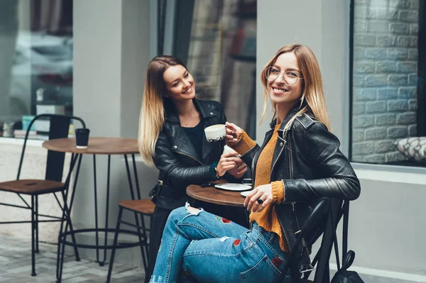 Δύο όμορφες νεαρές γυναίκες σε ρούχα της μόδας έχοντας υπόλοιπο μιλάμε και να πίνει καφέ στο υπαίθριο εστιατόριο. — Φωτογραφία Αρχείου