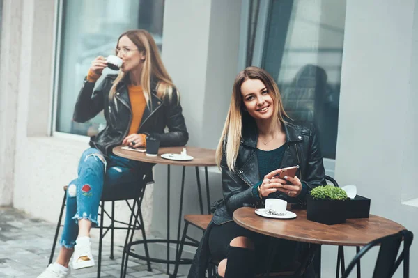 Veselá mladá žena sedící venkovní pití kávy s úsměvem a držení telefonu v ruce s krásnou módní dívka na pozadí. — Stock fotografie