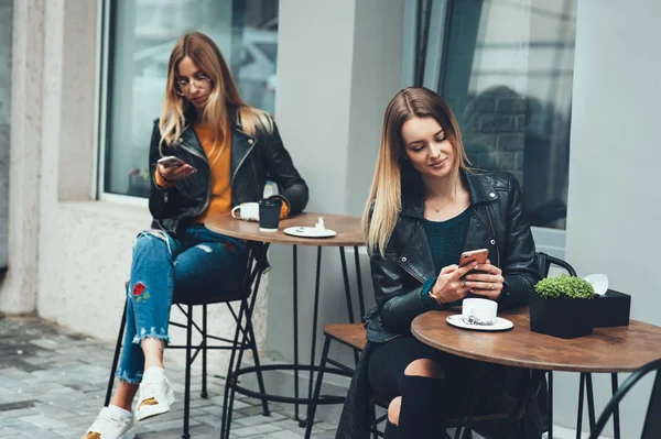 Δύο όμορφες νεαρές γυναίκες στο μοντέρνο φοράει συνεδρίαση υπαίθρια καφέ και χρησιμοποιώντας smartphones πίνοντας τον καφέ. Επιχειρηματικό σας μήνυμα. — Φωτογραφία Αρχείου