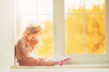 Sevimli küçük çocuk kız eve zevk gülümseyen iskambil büyük cam kenarında oturan pijama içinde. Penceresi Cacao Kupası ayakta. Sonbahar sezonu iç aile Tekstil Sanayi tatil insanlar kavramı