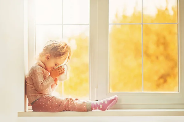 Милая маленькая девочка в пижаме сидит у большого окна и улыбается, наслаждаясь домом. Чашка какао стоит на окне. Осень Сезон интерьера Семейная текстильная промышленность Holiday People concept — стоковое фото