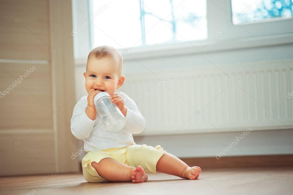 Little cute baby girl sitting in room on floor drinking water from bottle and smiling. Happy infant. Family people indoor Interior concepts. Childhood best time