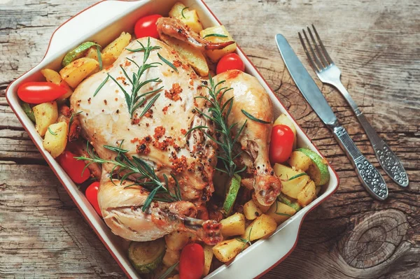 Roasted whole chicken stuffed with vegetables, tomatoes potato pepper and rosemary on vintage napkin wooden table background with fork and knife set. Top view, copy space, holiday food concept — Stock Photo, Image