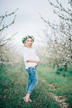Bahar geliyor! Güzel genç neşeli hamile kadında çelenk çiçek bahar ağacı bahçede yürürken göbek dokunmadan kafasına. Güzellik insanlar yaşam tarzı kavramları. Mutlu anne bebek için bekliyor!