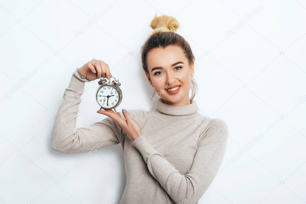 Portrait of beautiful young cheerful blonde woman with hair bun in sweater holding alarm clock and show emotions while standing over solated white background. Lifestyle Fashion Beauty Business Student