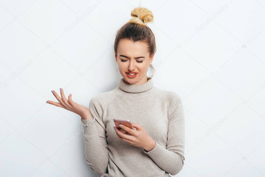 Horizontal portrait of displeased woman has indignant expression while holding smartphone, frowns eyebrows, can`t understand something, isolated on white background. Discontent emotion on face girl