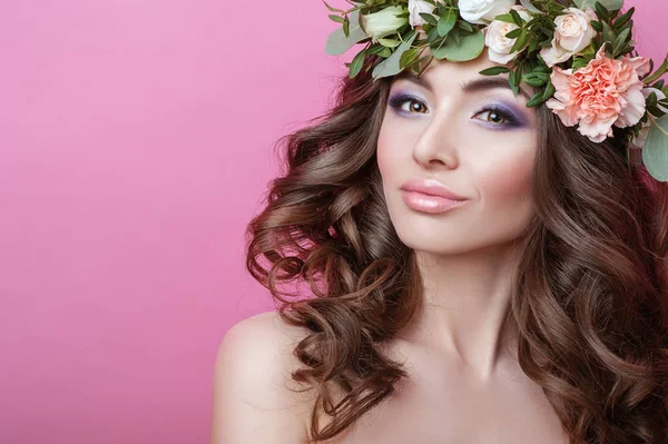 Beautiful young woman with curly hair and flower wreath on her head on pink background Beauty girl with flowers hairstyle Perfect makeup Beauty fashion People Skin