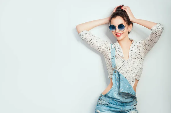 Lets dance! Young beautiful cheerful woman with hair bun in sunglasses wearing in jeans shorts dancing and posing over white wall. Good mood. Hands up! People Emotions Beauty Fashion Lifestyle concepts