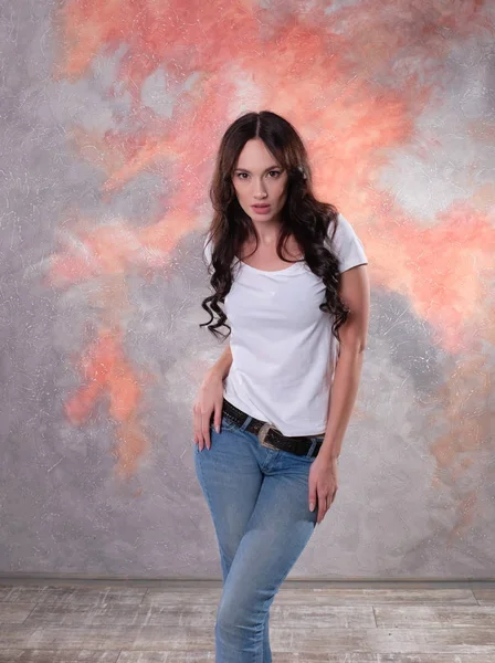 Mooie brunette vrouw in een denim jeans en witte T-shirt staande in sexy pose — Stockfoto