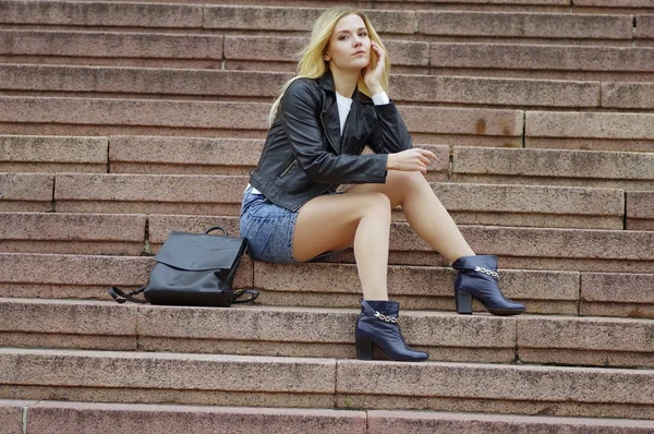 Uma menina bonita em uma jaqueta preta está sentada nas escadas . — Fotografia de Stock