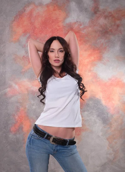 Vacker brunett kvinna i jeans och en vit T-shirt, stående i en sexig pose. — Stockfoto
