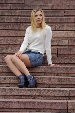 Güzel bir kız bir kot etek ve beyaz örgü kazak içinde belgili tanımlık merdiven üstünde oturur.