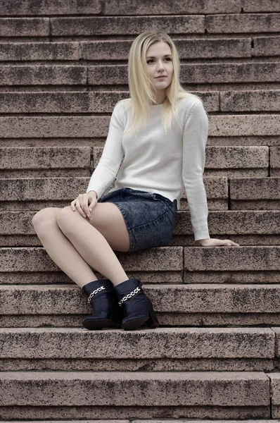 En vacker flicka i en denim kjol och en vit stickad tröja sitter på trappan. — Stockfoto