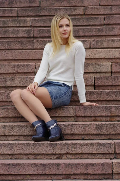 階段に座っているデニム スカートと白いニットのセーターの美しい女の子. — ストック写真