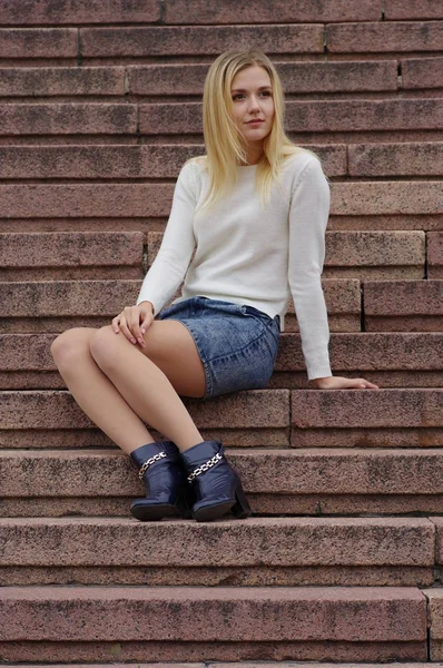 Krásná dívka v džínové sukně a bílý pletený svetr sedí na schodech. — Stock fotografie