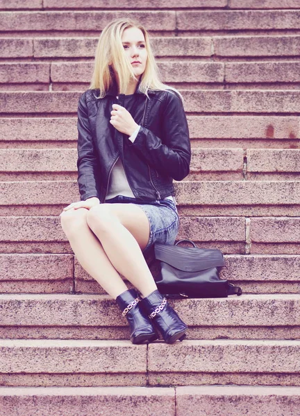 Una hermosa chica con una chaqueta negra y falda de mezclilla está sentada en las escaleras . — Foto de Stock
