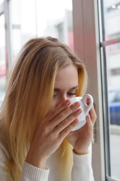 Retrato de una chica en un suéter de punto blanco bebiendo té en un café . — Foto de Stock