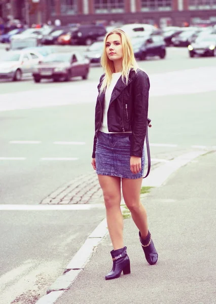 Una ragazza bionda dai capelli lunghi in gonna jeans e cappotto di pelle in piedi in una strada . — Foto Stock