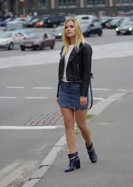 Seorang gadis rambut panjang pirang dengan rok jins dan jaket kulit berdiri di jalan . — Stok Foto