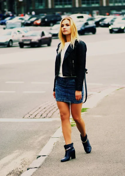 Una chica rubia de pelo largo con falda vaquera y abrigo de cuero de pie en una calle . — Foto de Stock