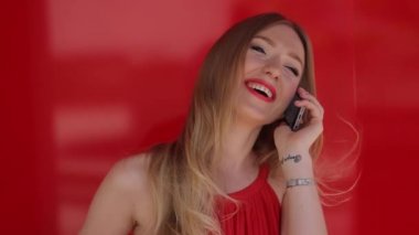 Birisine kırmızı arka plan ile telefonda konuşurken Kırmızı elbiseli uzun saygısız saçlı genç, seksi, çekici, güzel, gülümseyen sarışın kadın,